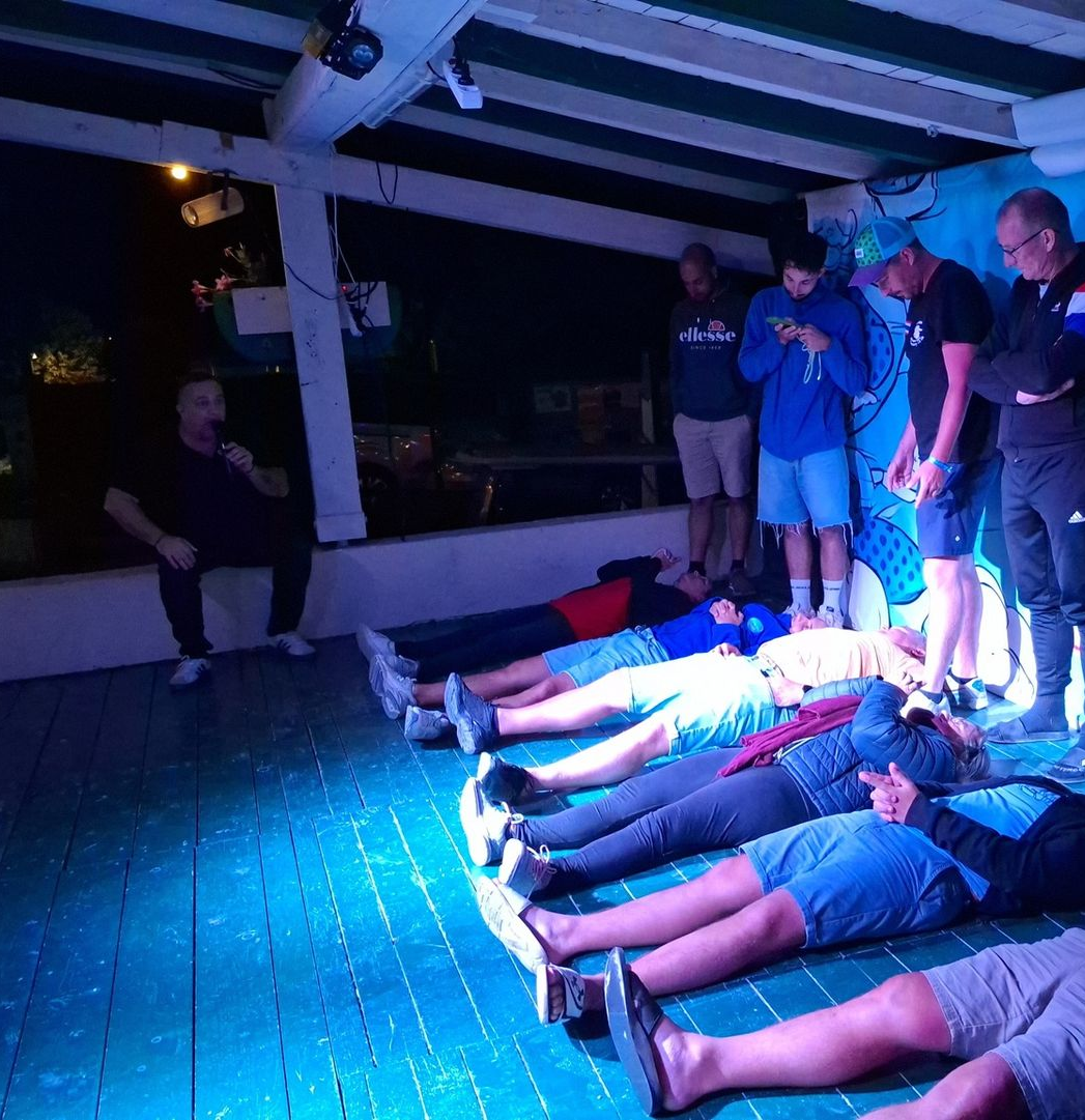 David Parrat Hypnotiseur s'adresse à des personnes allongées sur une estrade. D'autres restent debout et regardent. Décor extérieur sombre, éclairé d'une lumière bleutée.