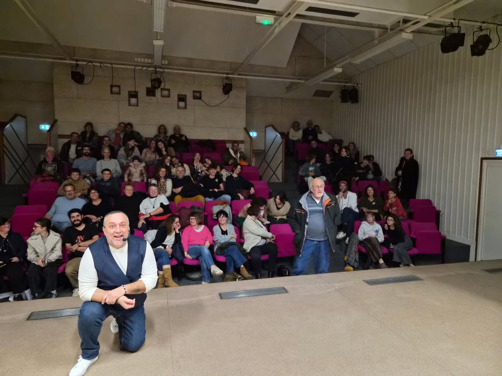 David Parrat Hypnotiseur s'agenouille devant le public d'un théâtre. Les gens sont assis en rangs serrés et le regardent ; l'homme sourit.