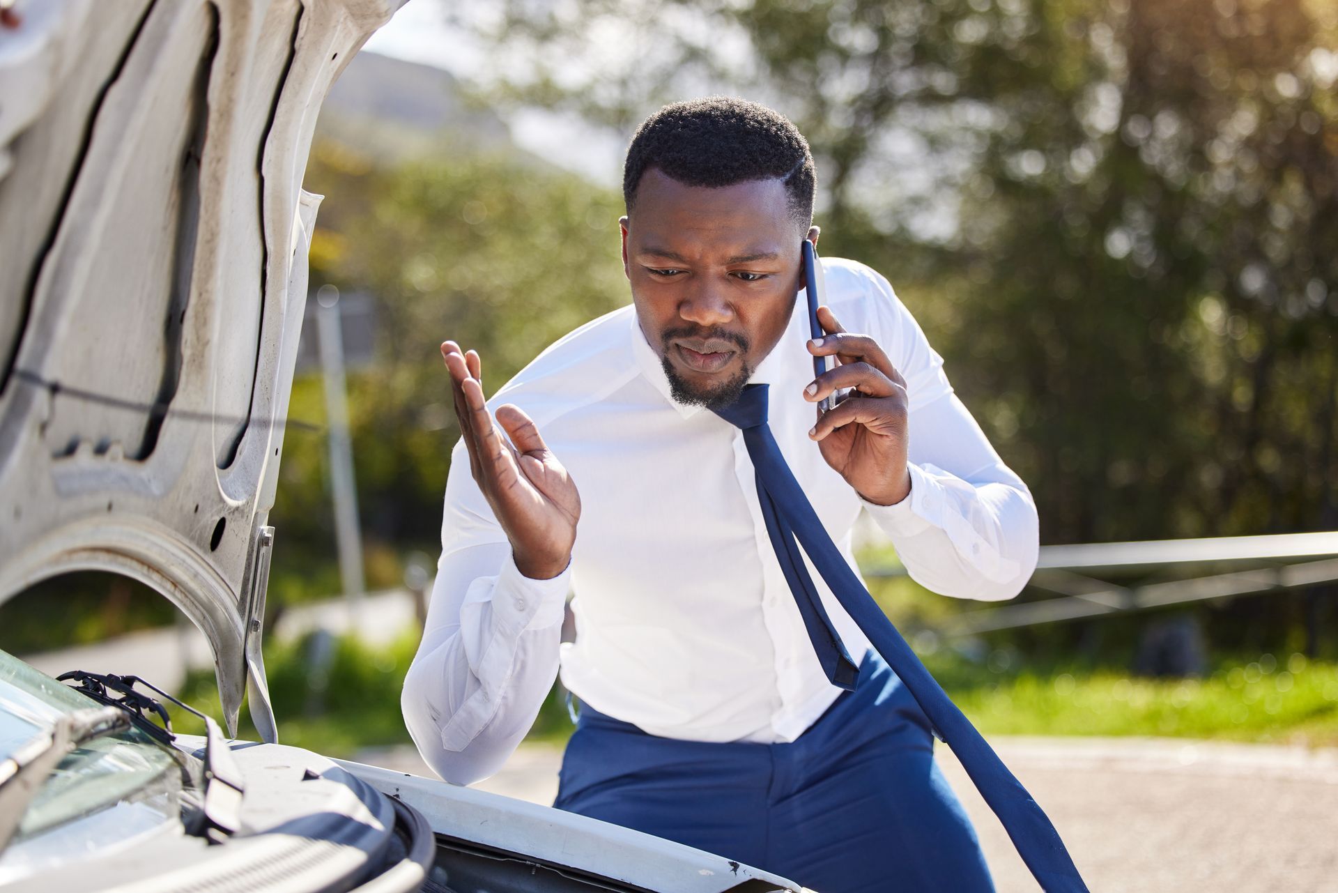 Un homme en chemise et cravate au téléphone regardant le moteur sous le capot