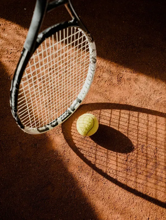 Raqueta de tenis proyectando una sombra sobre una pelota de tenis en una cancha de arcilla.
