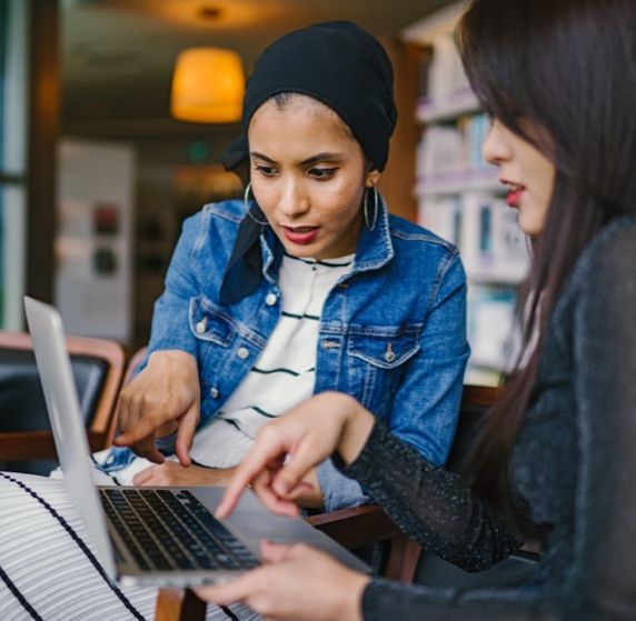 Dos mujeres están sentadas en una mesa mirando una computadora portátil.