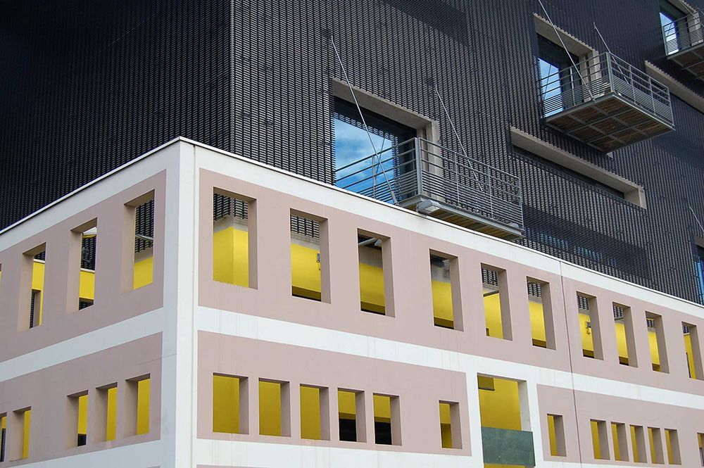 Exterior de um edifício moderno com estrutura rosa claro e janelas com fundo amarelo, sob uma fachada de malha metálical.