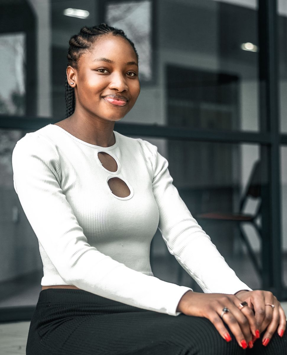 Frau mit geflochtenem Haar, lächelnd, trägt ein weißes, ausgeschnittenes Oberteil und eine schwarze Hose und sitzt vor einem Fenster.