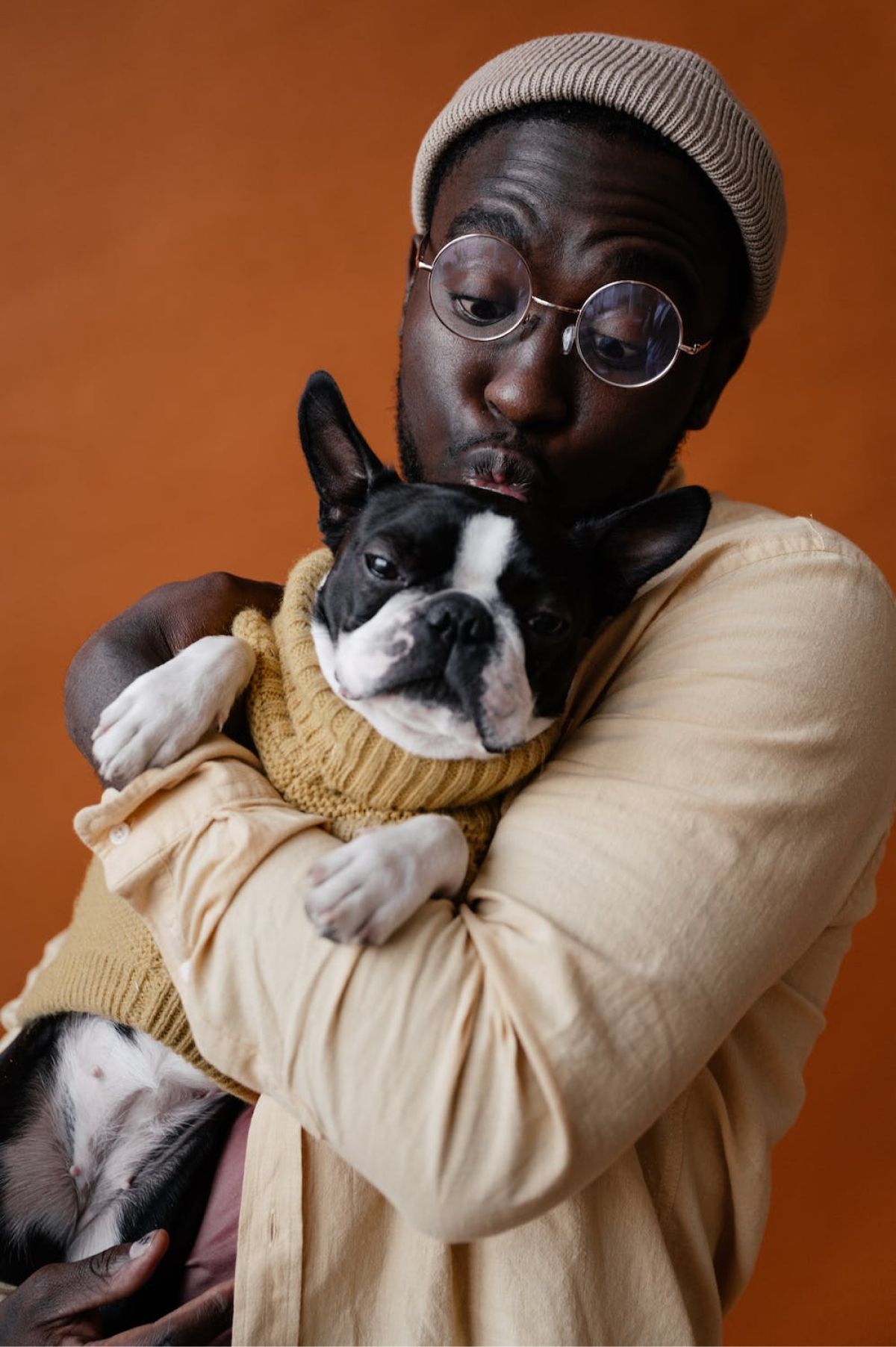 Een persoon met een beige pet en een beige overhemd omhelst een Bostonterriër die een bijpassende gele trui draagt, terwijl hij zijn lippen tuurt.