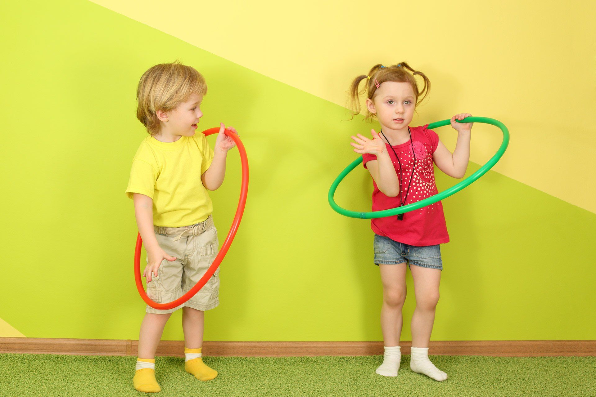 Ein Junge und ein Mädchen halten Hula-Hoop-Reifen und stehen vor einer grün-gelben Wand.