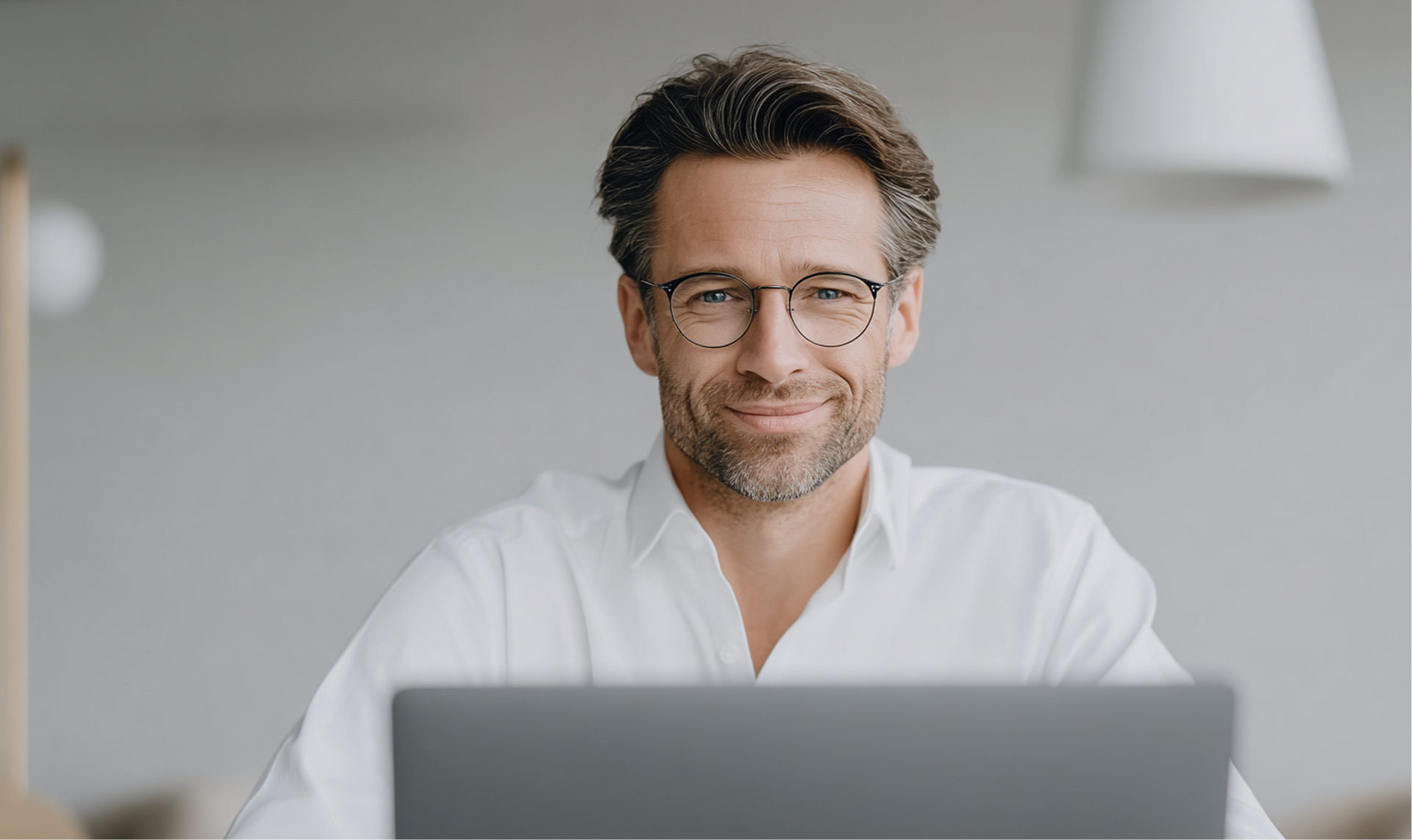 Mann mit Brille lächelt hinter einem Laptop in einem hellen Büro