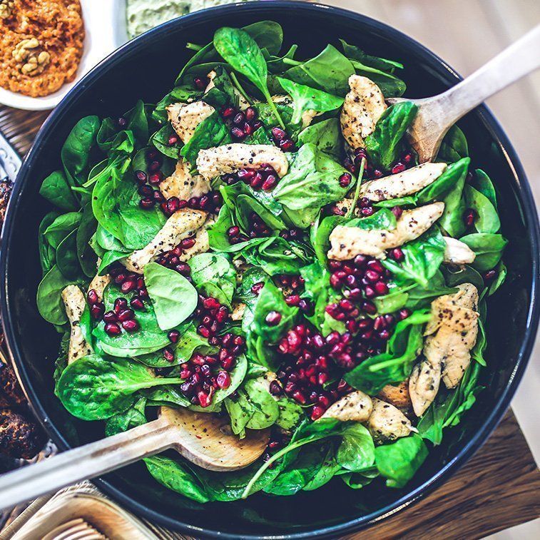 A salad with spinach , chicken , pomegranate seeds and a spoon in a bowl.