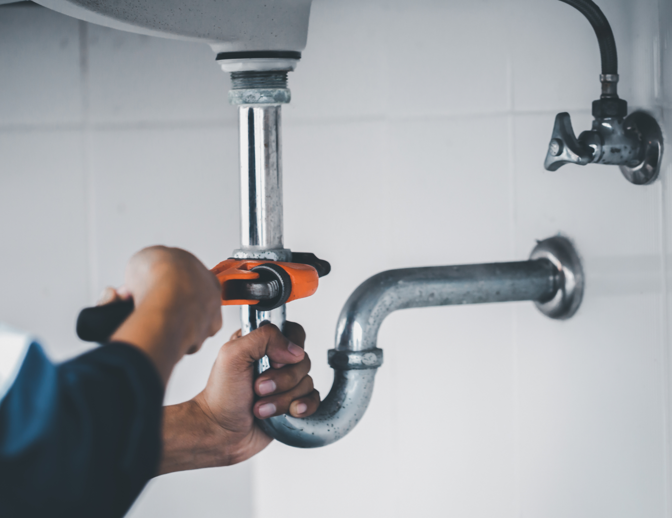 Un fontanero usa una llave inglesa para reparar una tubería curva debajo de un lavabo. El entorno es un baño.
