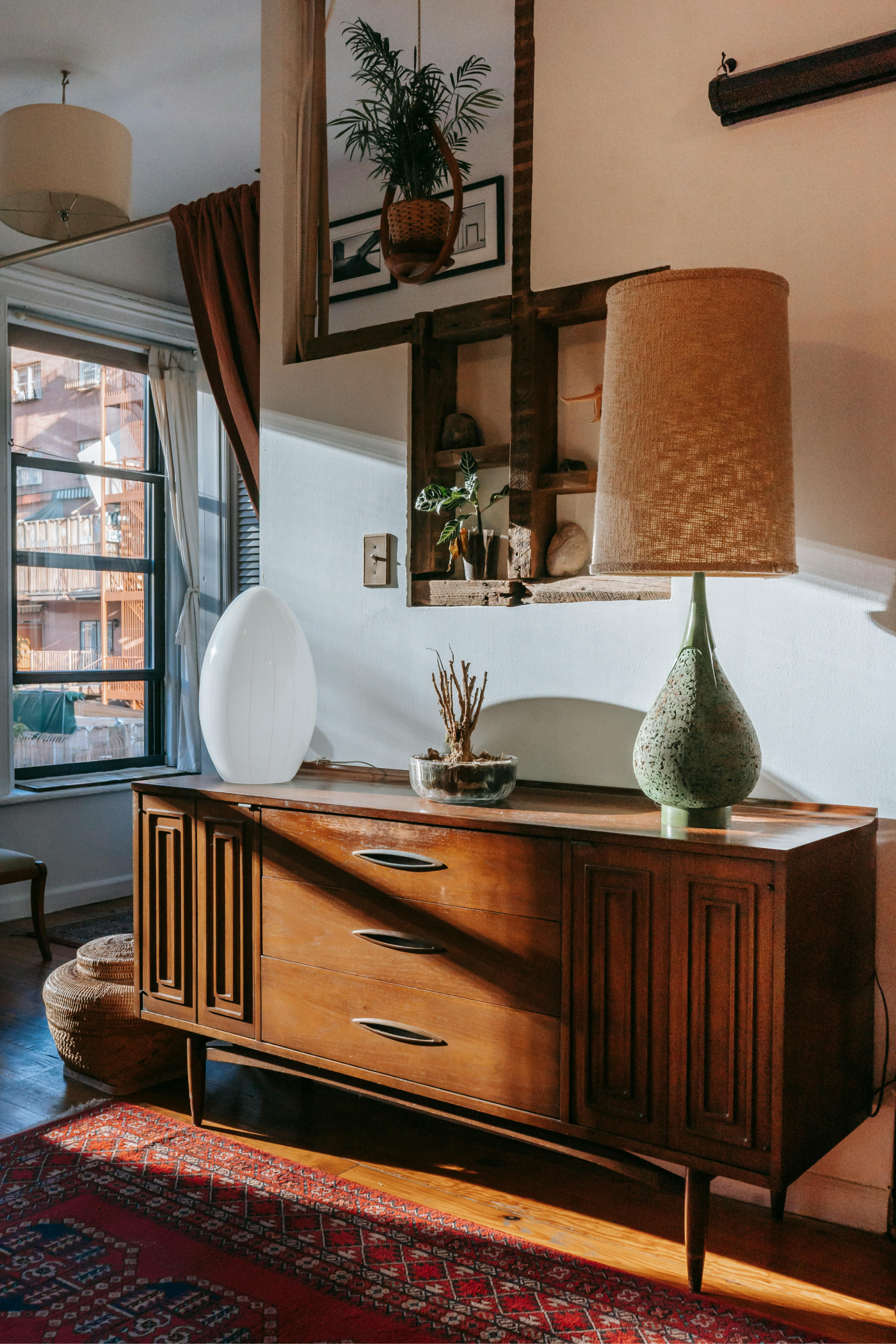 A mid-century modern wooden cabinet with a lamp and decor sits in a room with a window and rug, bathed in sunlight.