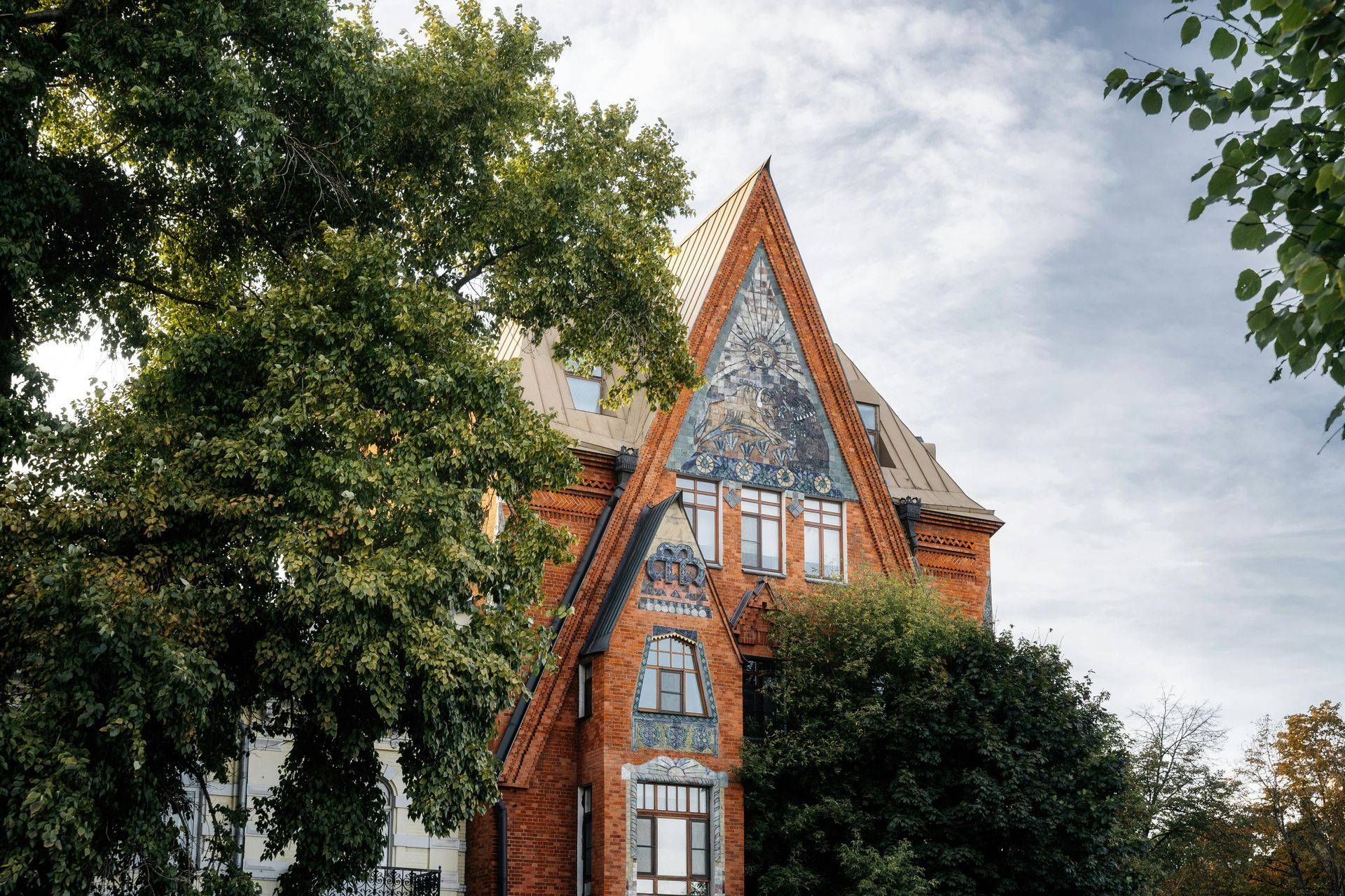 Red brick building with intricate details under a cloudy sky, framed by green trees.