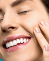 Una mujer de piel radiante sonríe, tocándose la mejilla. Labios rosados, ojos cerrados.