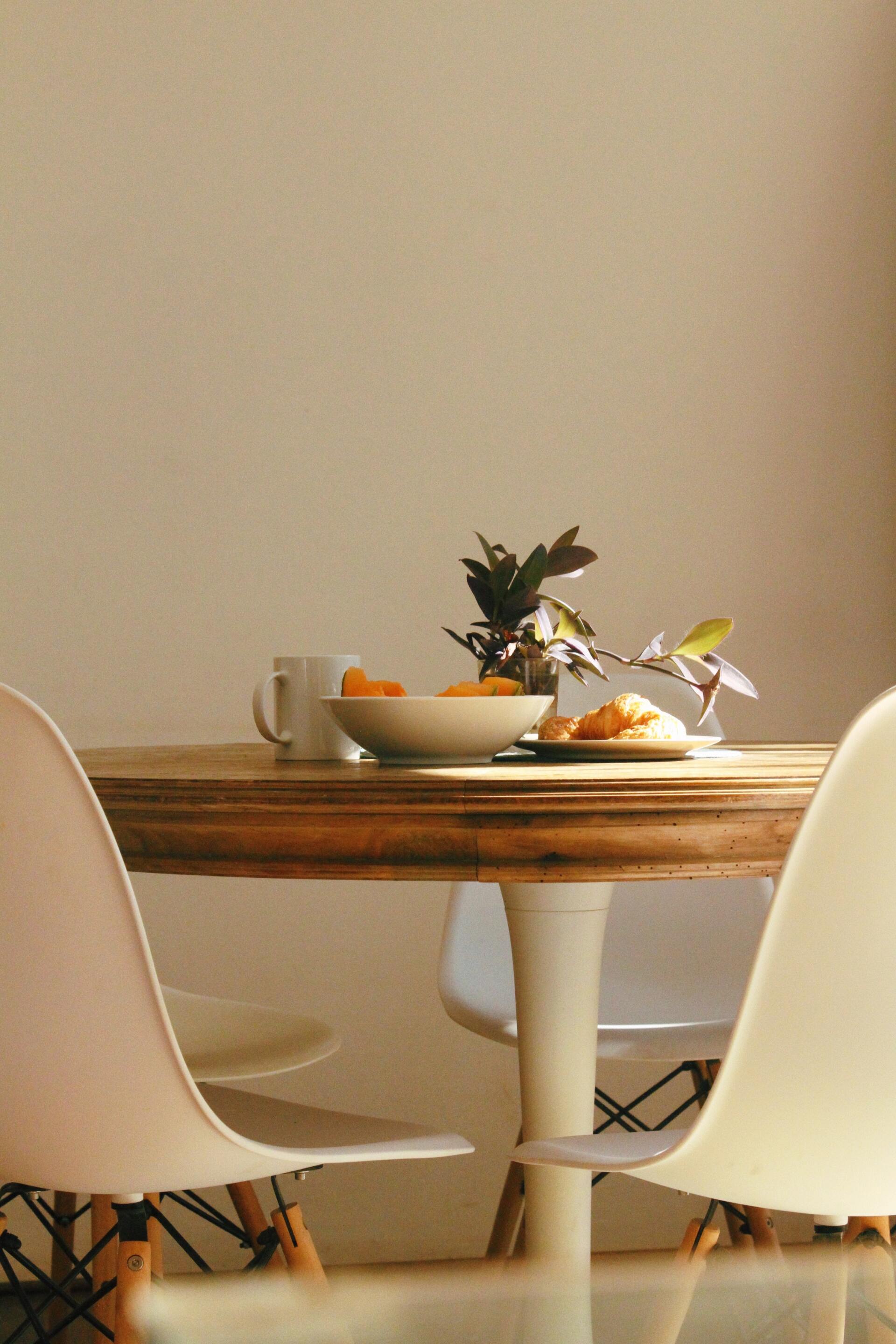 Table à manger avec une corbeille de fruits et des plantes entre deux chaises blanches contre un mur beige