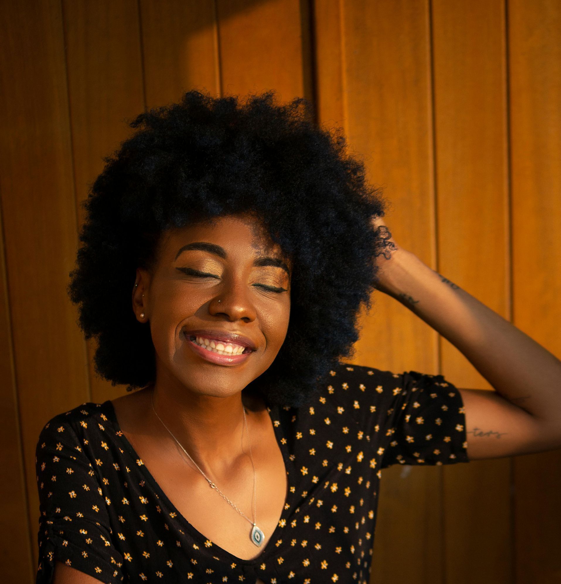 Femme souriante aux cheveux bouclés, posant contre un mur en bois, vêtue d'une robe noire à pois dorés.