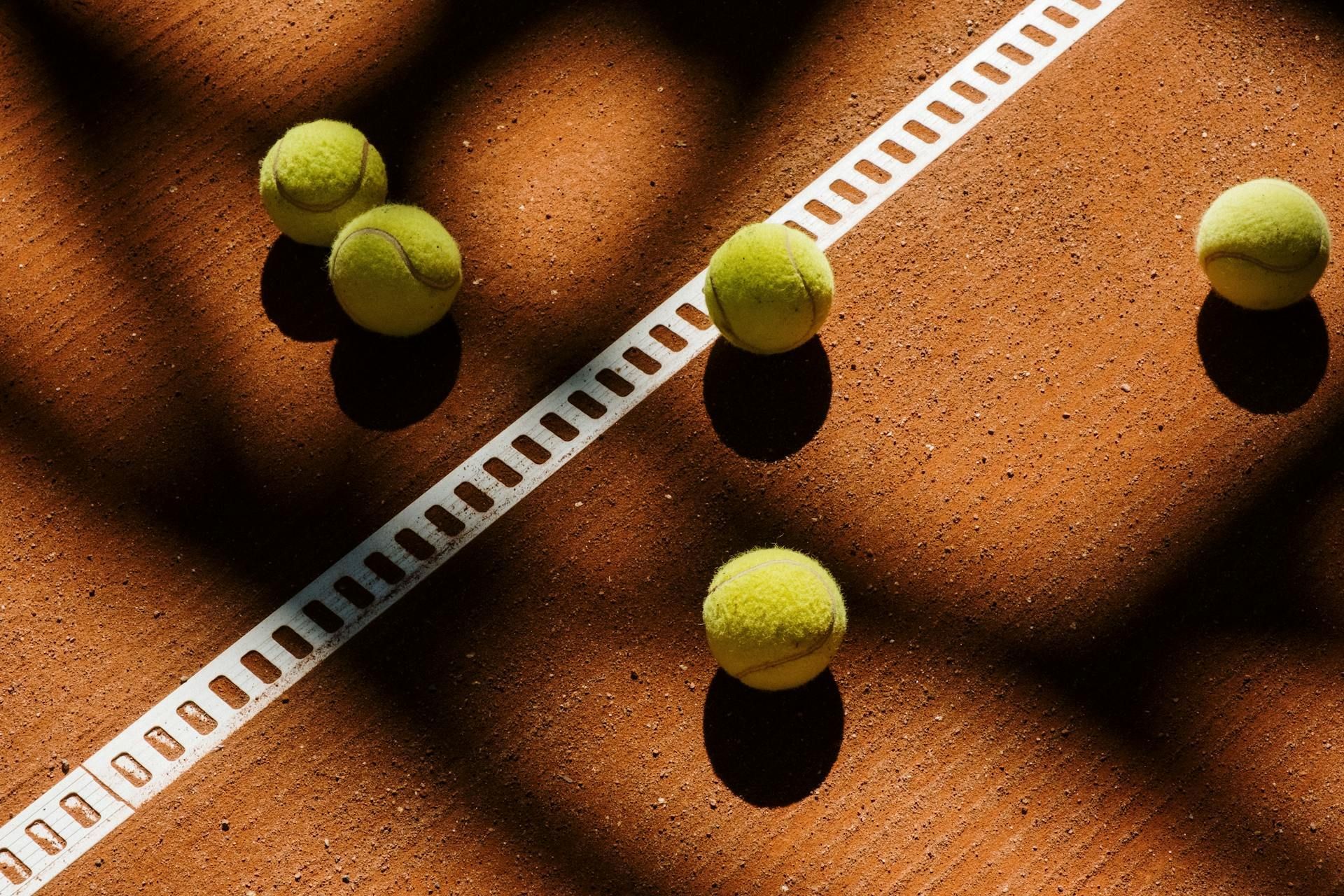 Vier gelbe Tennisbälle auf einem Sandplatz mit weißer Linie und Netzschatten