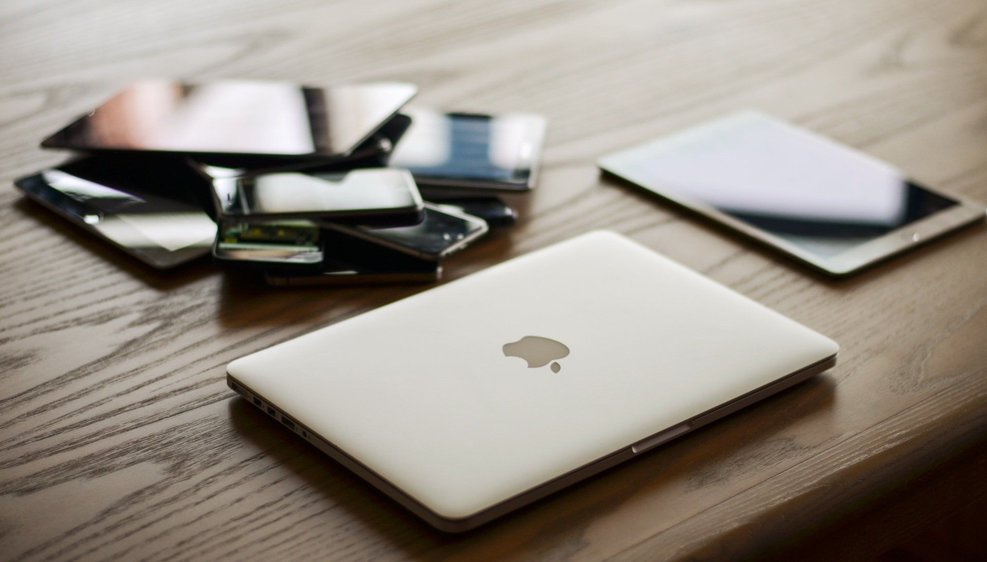 Auf einem Holztisch steht ein Apple-Laptop neben einem Stapel Handys und einem Tablet.