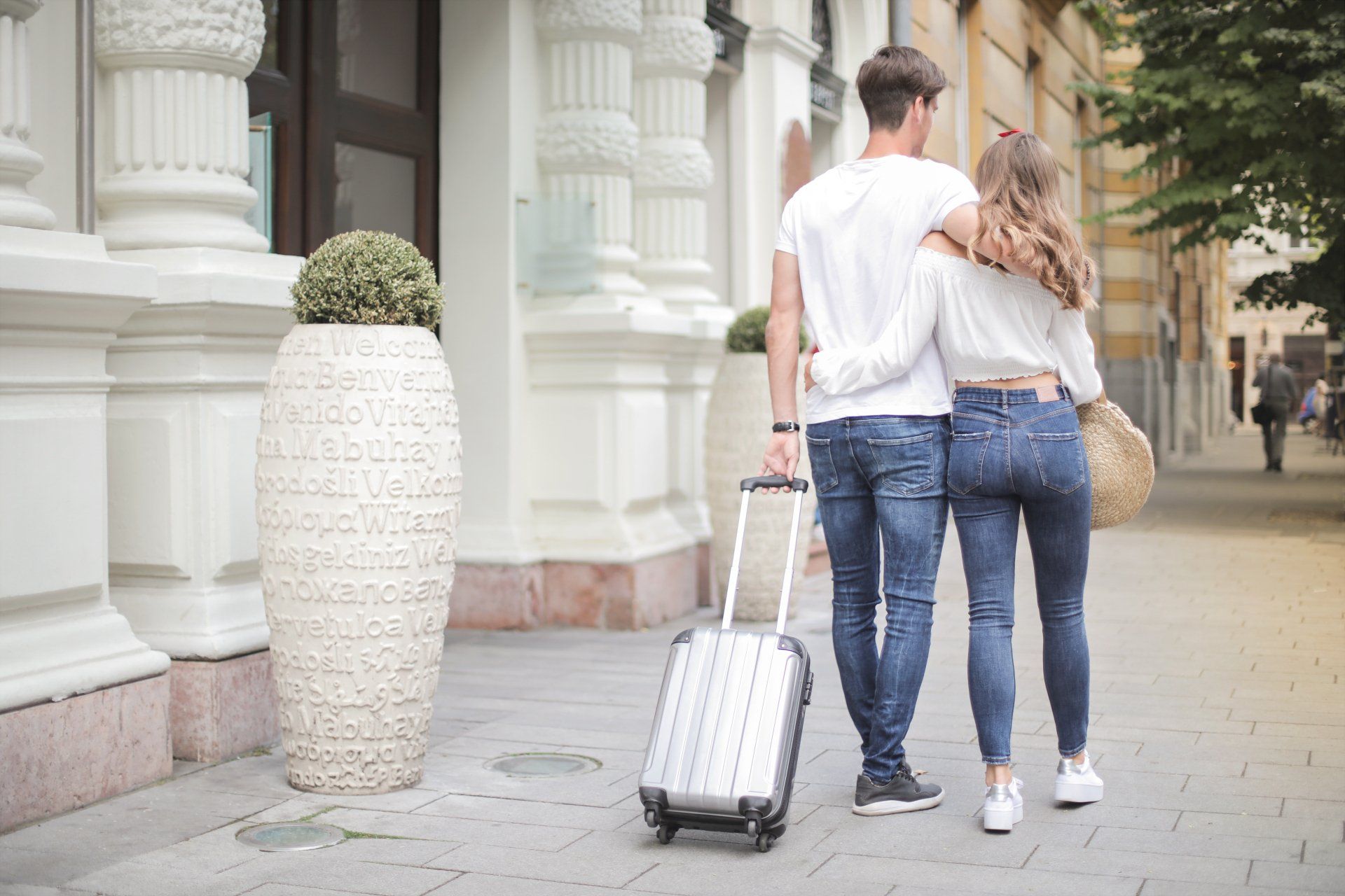 Couple walking, embracing, with luggage in front of a building. They are in jeans and white tops.