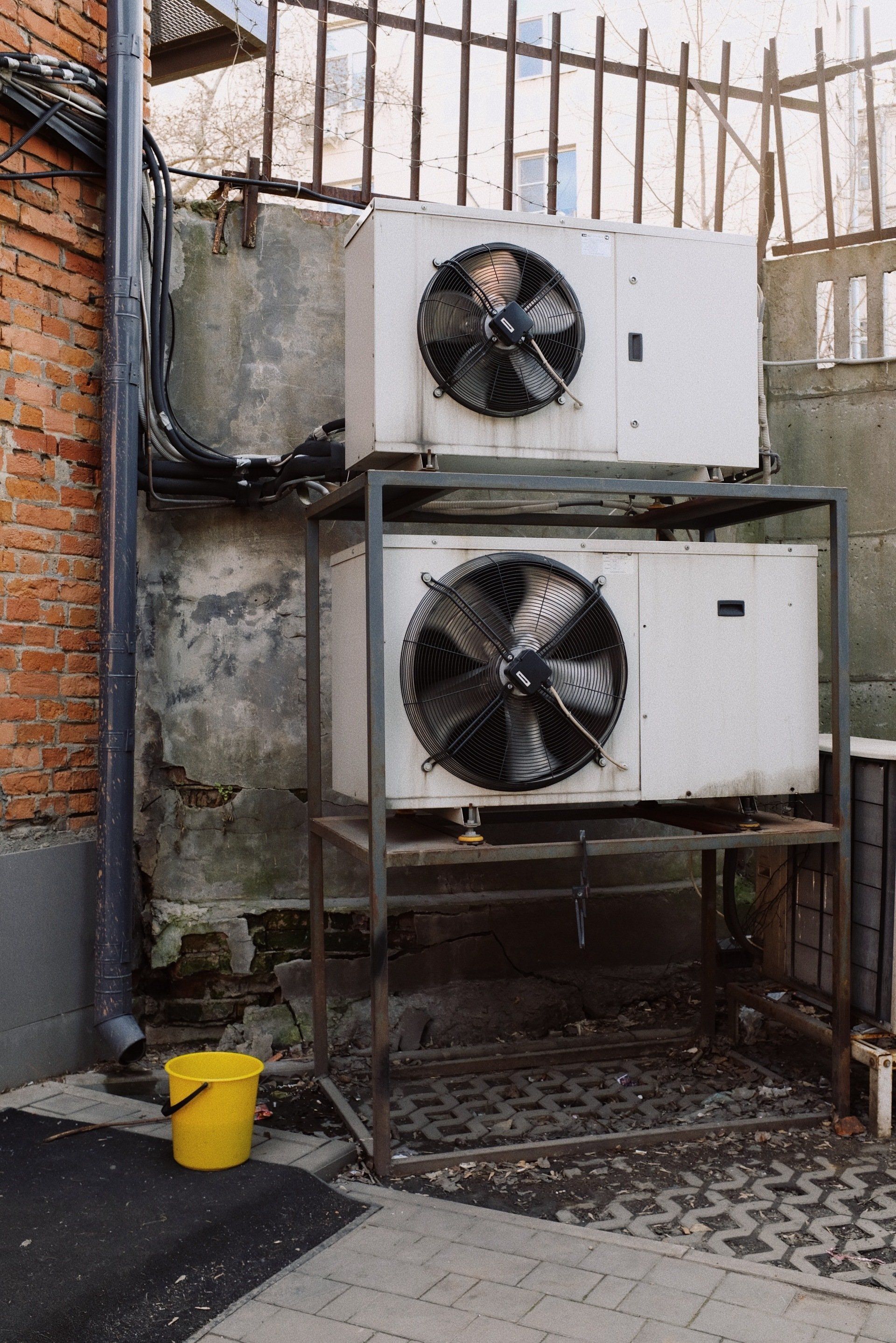 Two air conditioners are stacked on top of each other next to a yellow bucket.