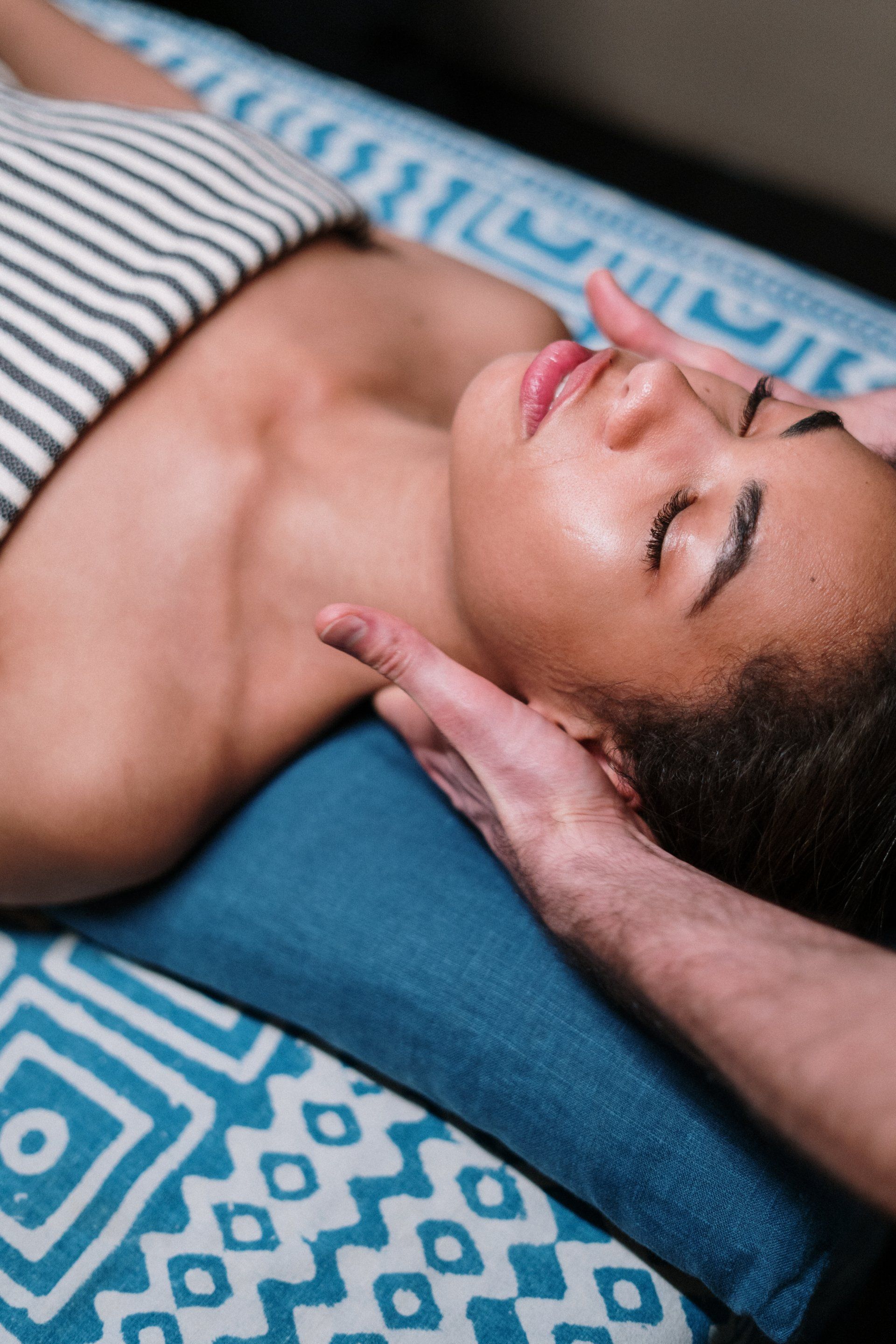 Person, die eine Gesichtsmassage erhält, liegt auf einem blauen Kissen, die Augen geschlossen, die Hände auf dem Kopf.