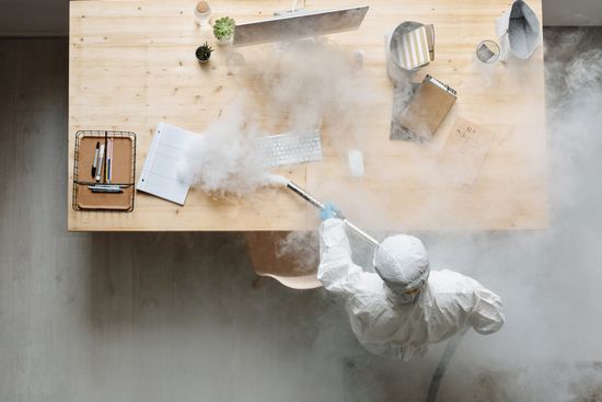 A man in a protective suit is disinfecting a desk with a spray bottle.