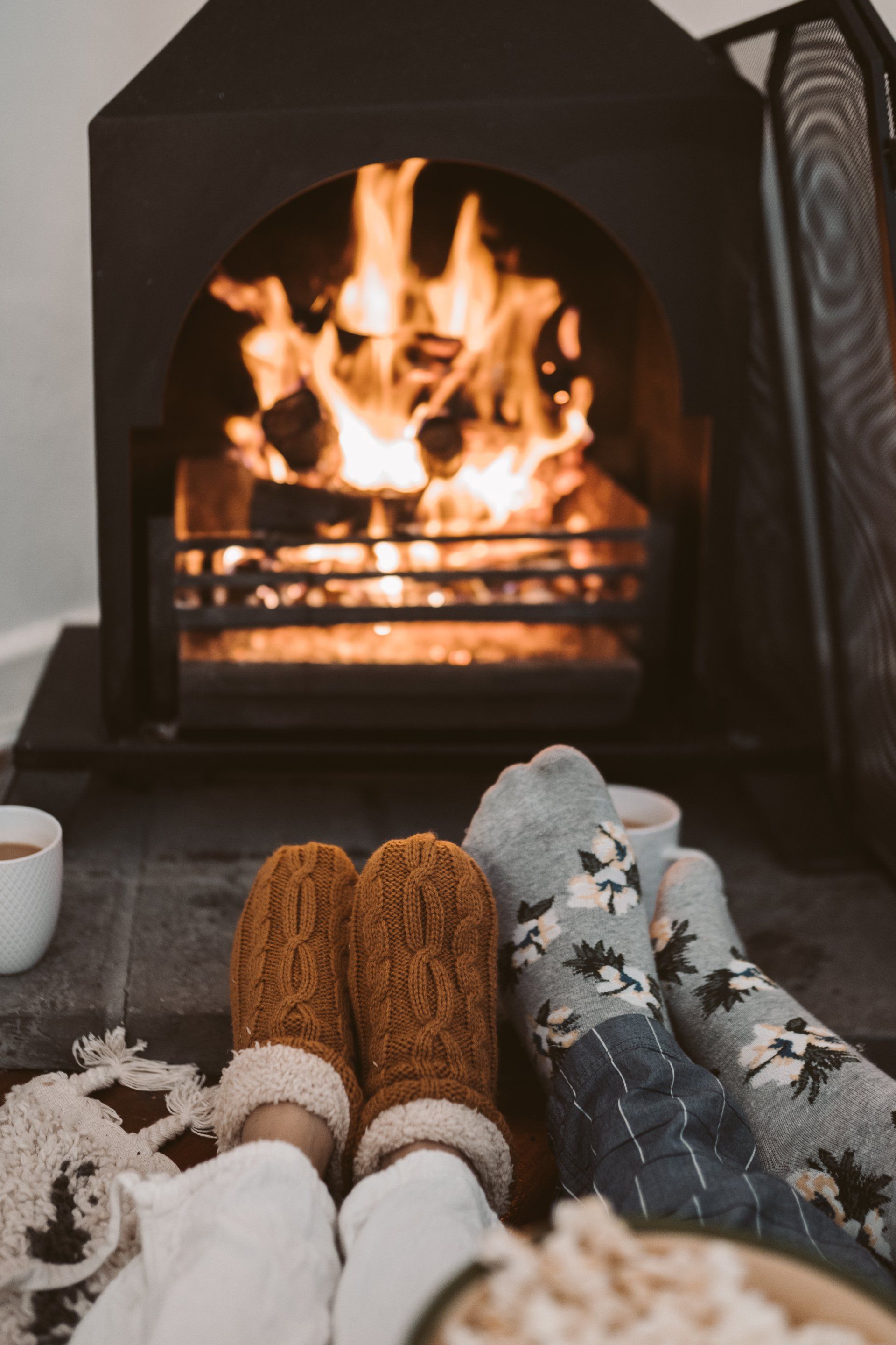 Füße in kuscheligen Socken und Fäustlingen vor einem Kamin mit brennenden Flammen.