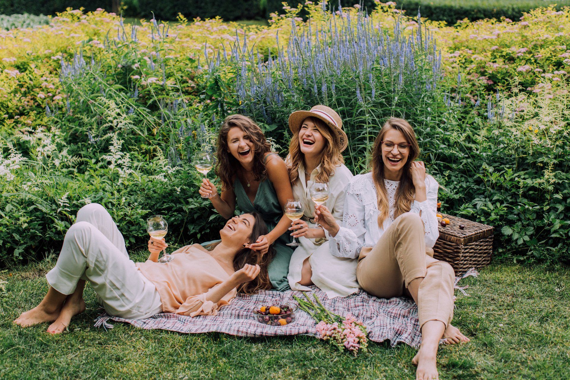 Vier lachende Frauen bei einem Picknick in einem Garten, halten Getränke in der Hand und lächeln.
