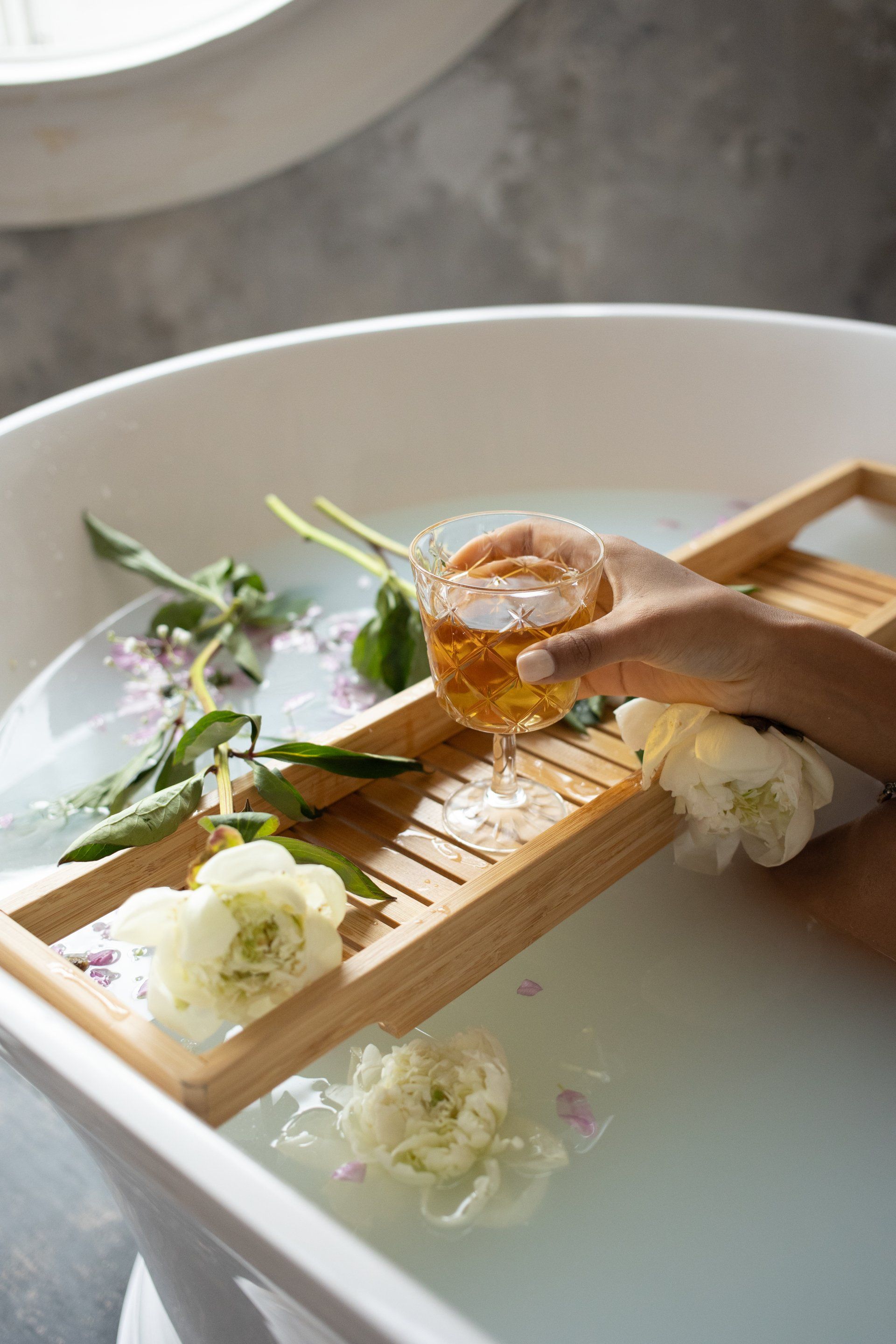 Hand hält ein Glas mit Flüssigkeit in einer Badewanne mit weißen Blumen und Blütenblättern auf einem Holztablett.