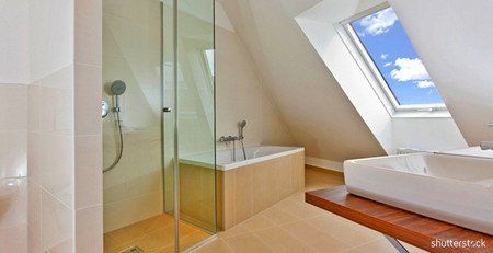 Salle de bains avec douche, baignoire, puits de lumière et lavabo blanc. Journée ensoleillée. Plafond mansardé.