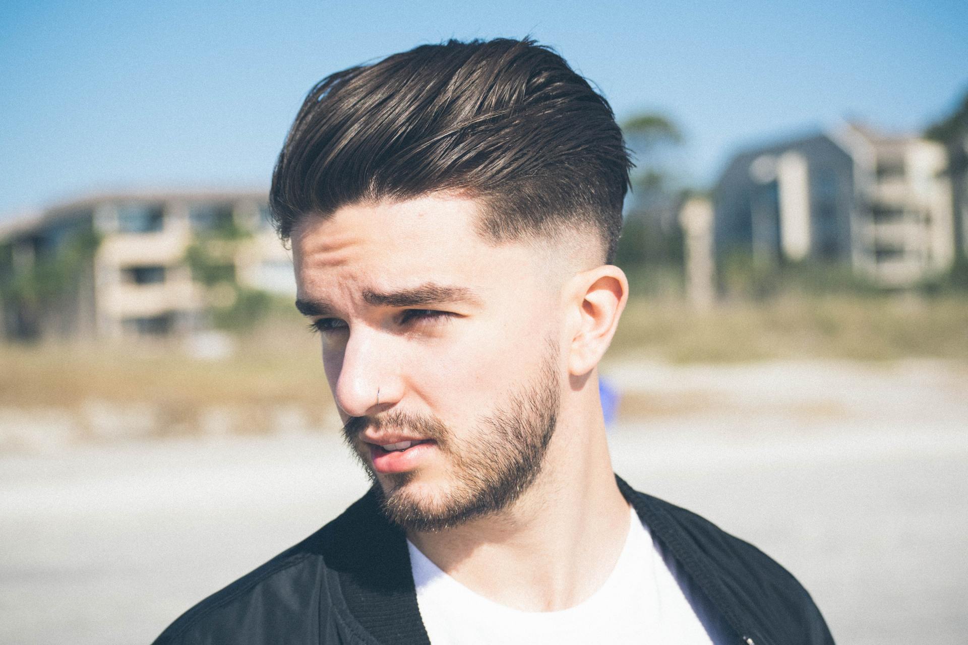 Un homme aux cheveux dégradés et à la barbe fournie regarde au loin sur une plage.